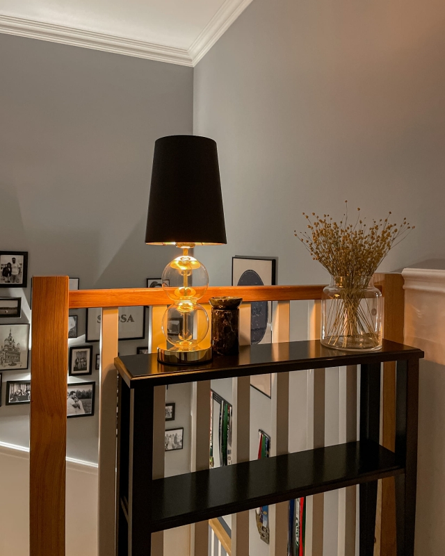 Złota lampka stołowa ze szklanymi elementami i czarnym abażurem Kaspa COCO w aranżacji na konsoli na klatce schodowej.