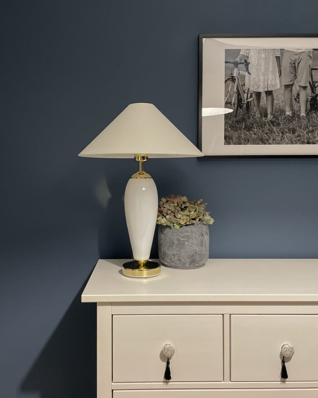 Biało-złota lampa stołowa REA z białym abażurem w kształcie stożka w aranżacji na komodzie.