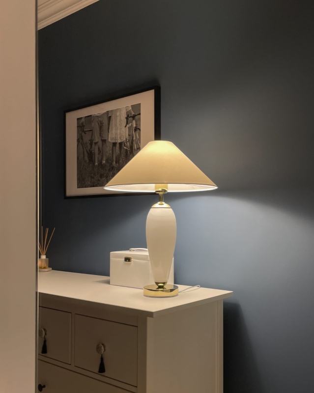 Biało-złota lampa stołowa REA z białym abażurem w kształcie stożka w aranżacji na komodzie.